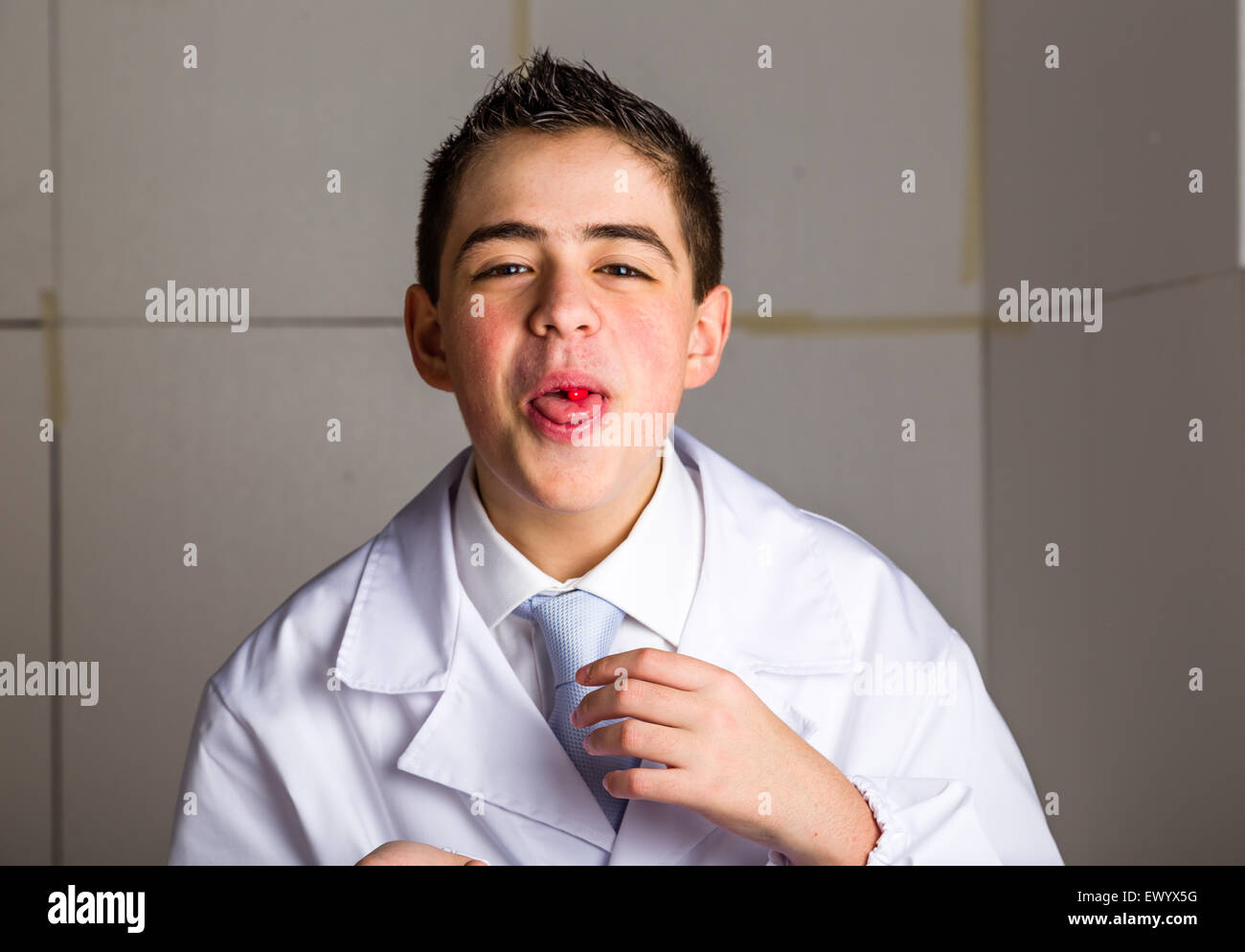 Boy dressed as a doctor in light blue tie and white coat helps to feel medicine more friendly: he is showing his tongue with a pill on it. His acne skin has not ben retouched Stock Photo