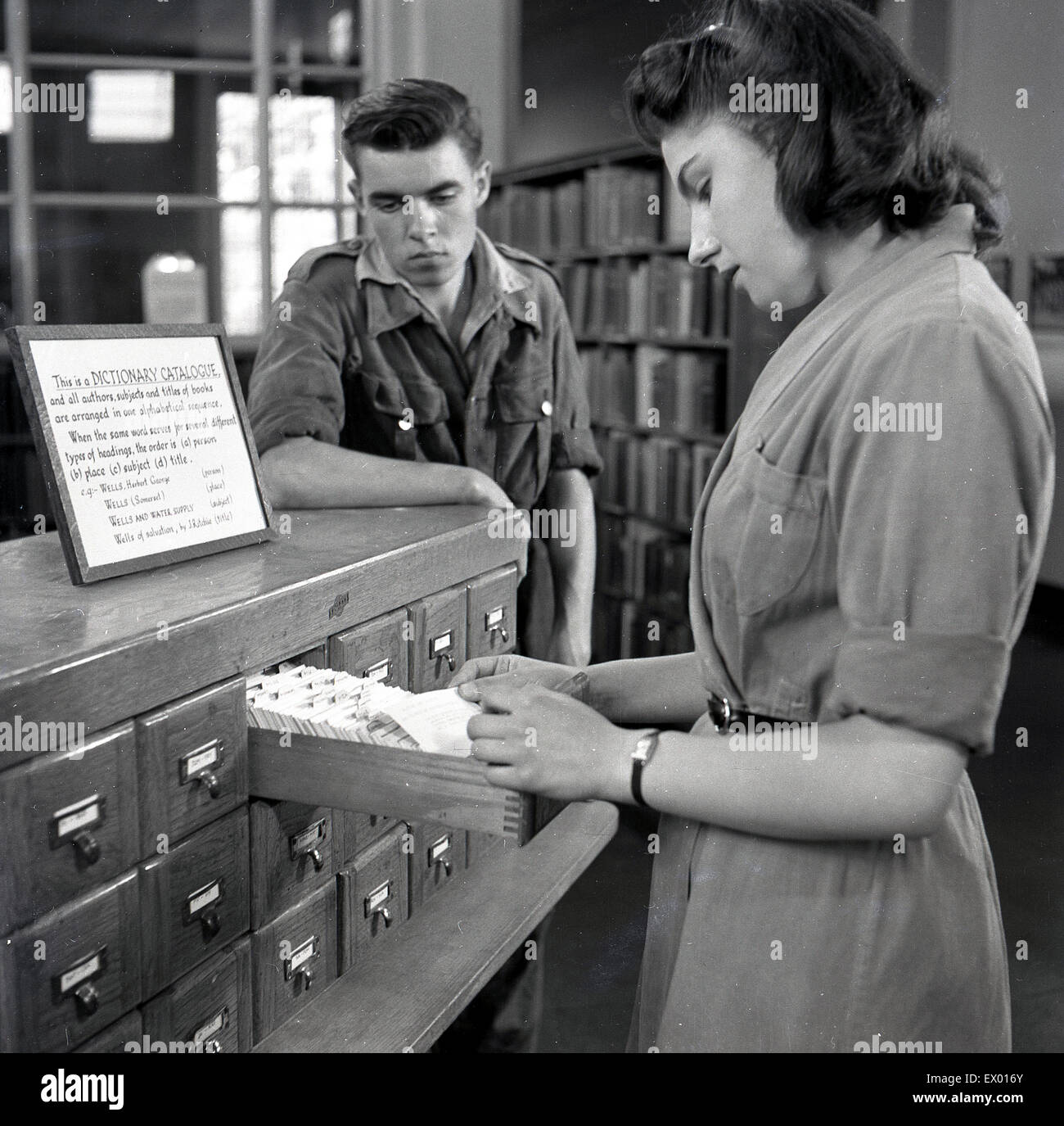 Historical, 1950s, young female librarian looks in a card index tray in search of a book a young man is looking for. Stock Photo
