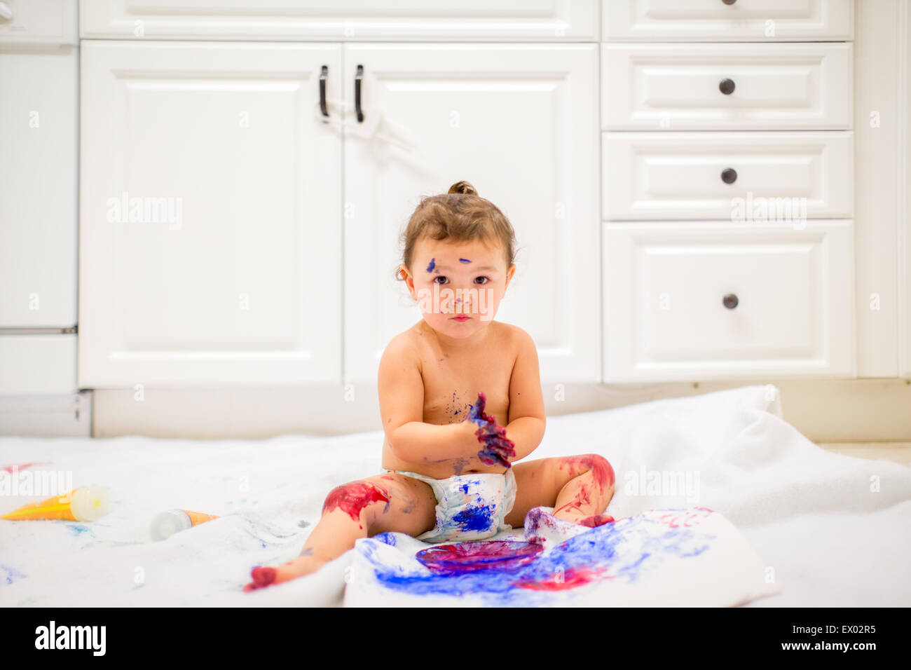 Baby girl dabbling with finger paints in white kitchen Stock Photo