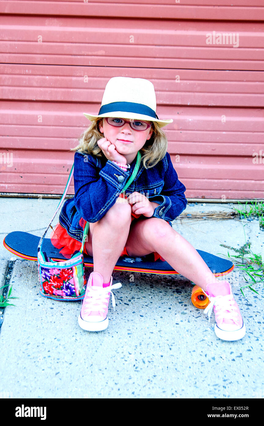 Portrait of young girl wearing tutu, denim jacket and hat, sitting on skateboard Stock Photo