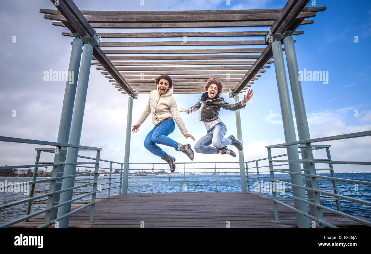 Two young women friends jumping in unison on sea pier Stock Photo