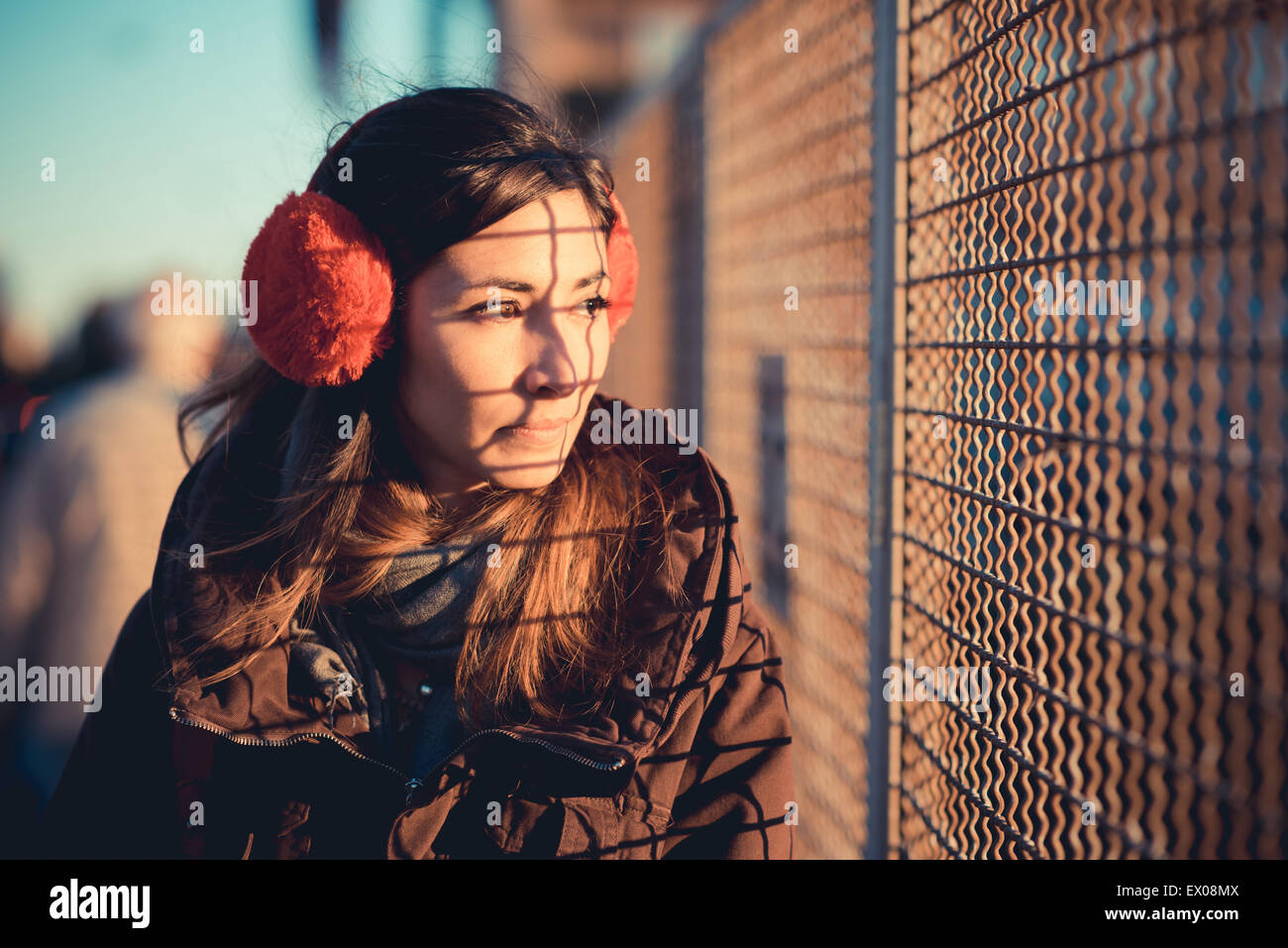 Mid adult woman wearing red ear muffs gazing through sunlit wire fence Stock Photo