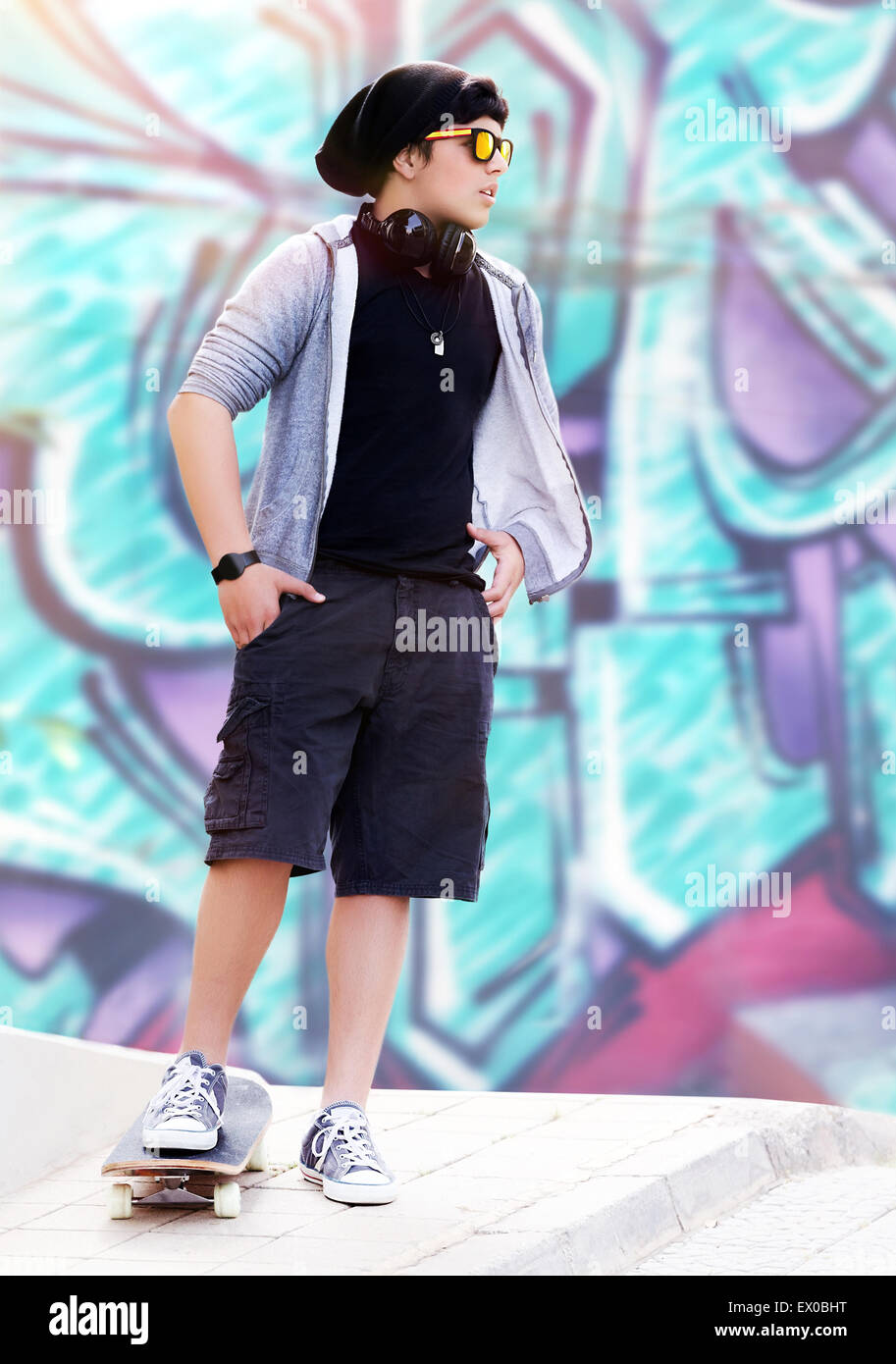 Stylish skater boy listening music standing outdoors on beautiful graffiti wall background, active teen life, hipster style Stock Photo