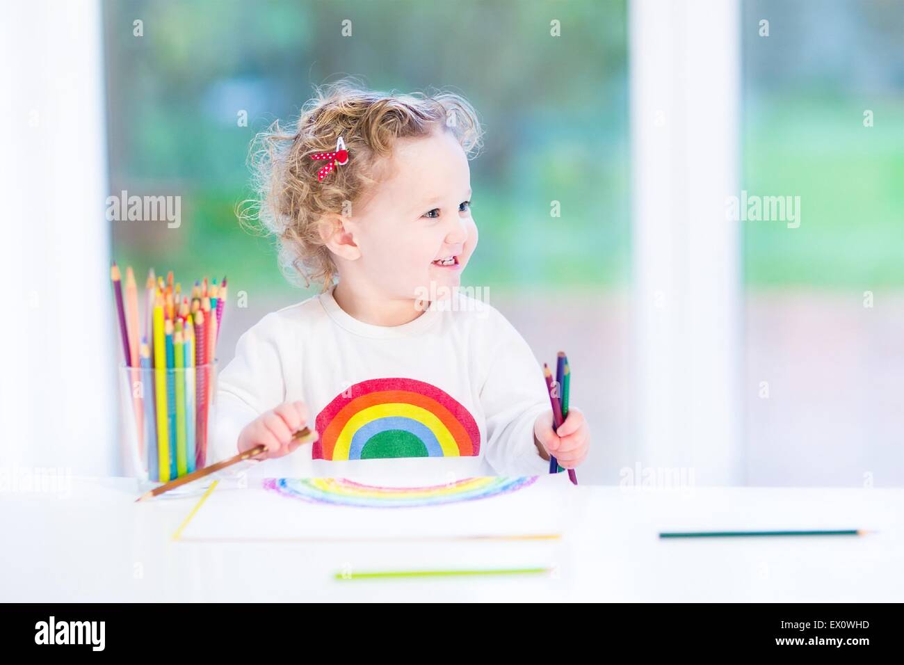 Funny smiling toddler girl drawing a rainbow with colorful pencils at a white desk sitting next to a big window into the garden Stock Photo