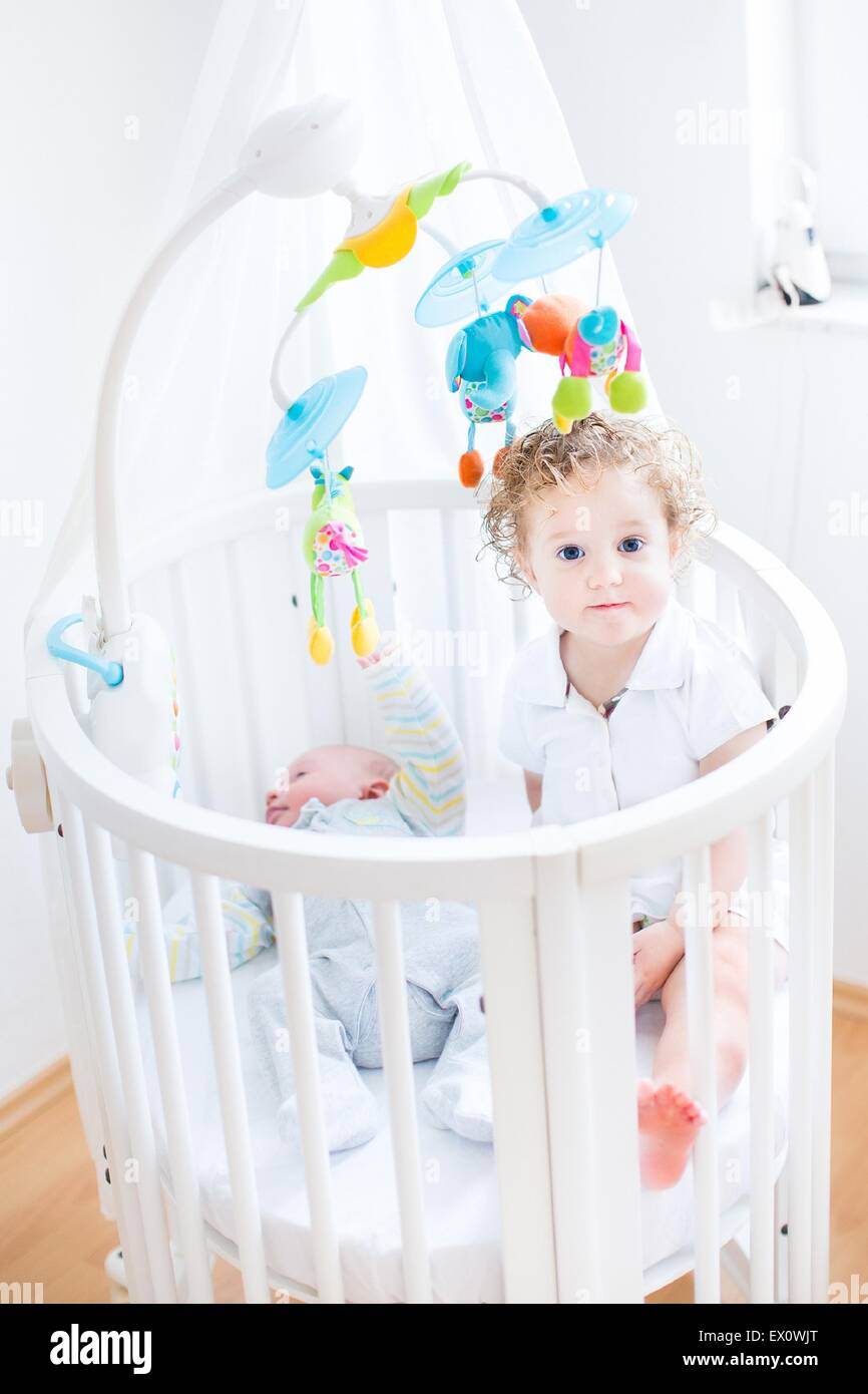 Cute funny toddler girl sitting in a crib of her her newborn baby