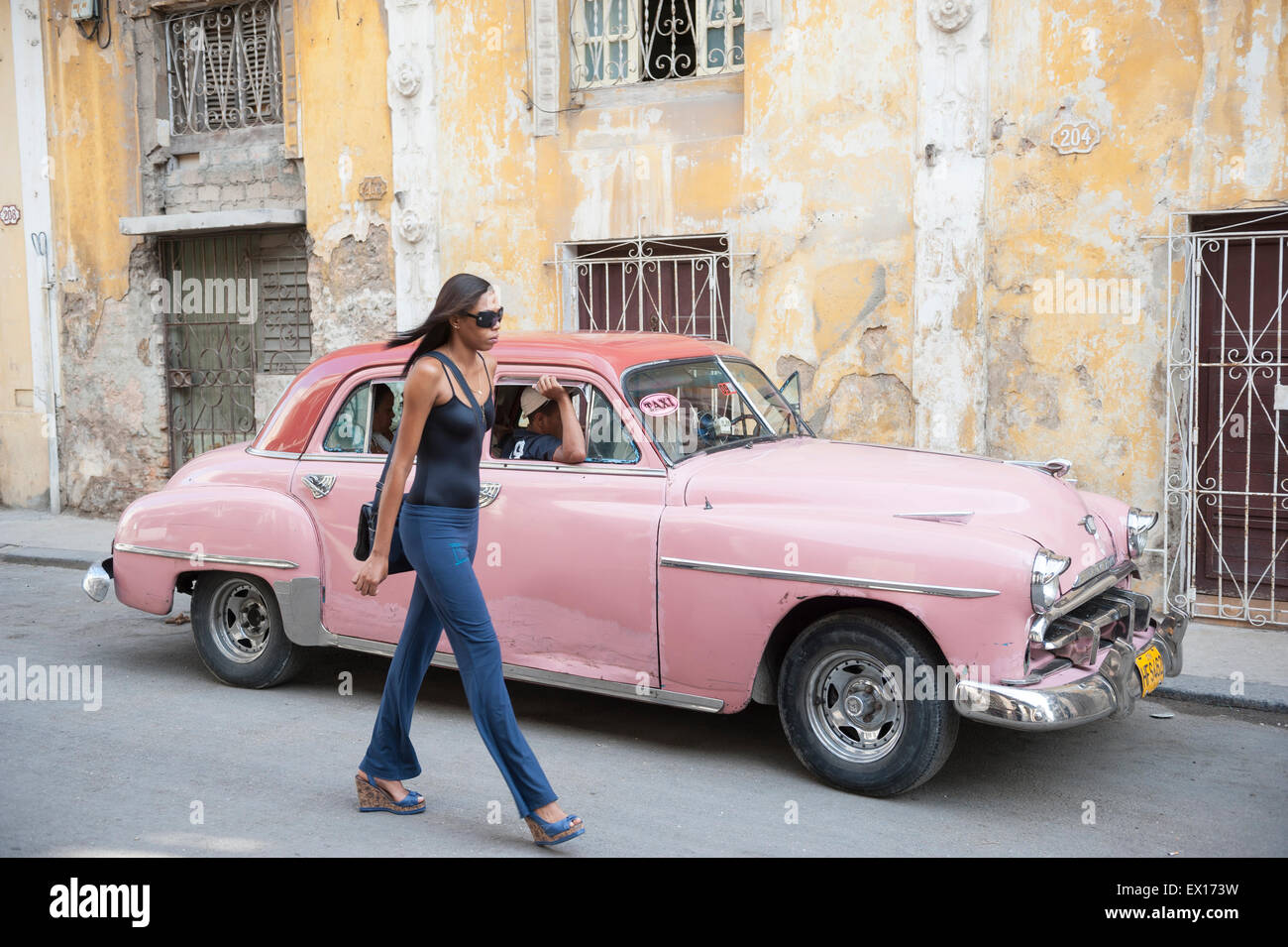HAVANA, CUBA - JUNE 13, 2011: Young Cuban woman walks beside vintage American taxi car in front of weathered colonial buildings. Stock Photo
