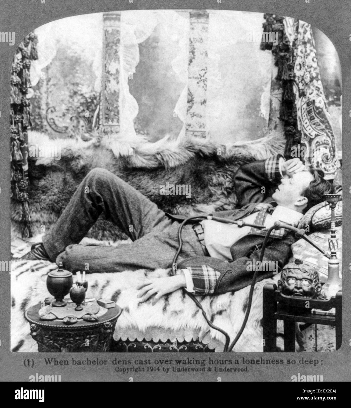 Romanticized studio portrait of opium use entitled 'When bachelor dens cast over waking hours a loneliness so deep.' Image shows a young man relaxing on a fur covered chaises lounge with a hookah on the left and various smoking equipment on the left. This romanticized view of carefree opium use started with De Quincey's popular 'Confessions of an English Opium-Eater' and endured until the 1920's. Image from a stereograph released by Underwood & Underwood in 1904. Stock Photo