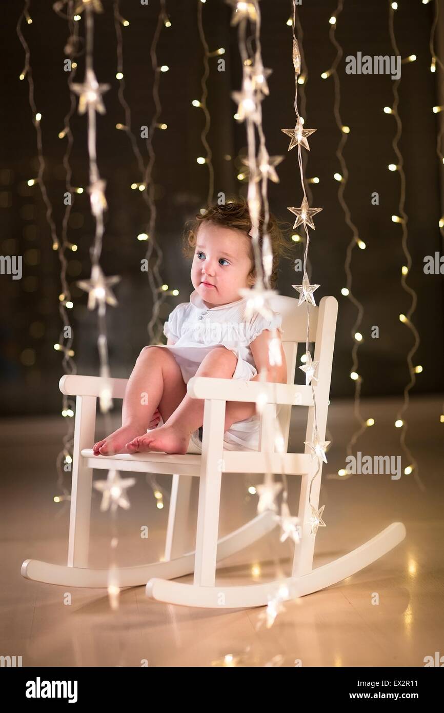 Adorable toddler girl in a white dress sitting in a white rocking chair in a dark room with Christmas lights Stock Photo