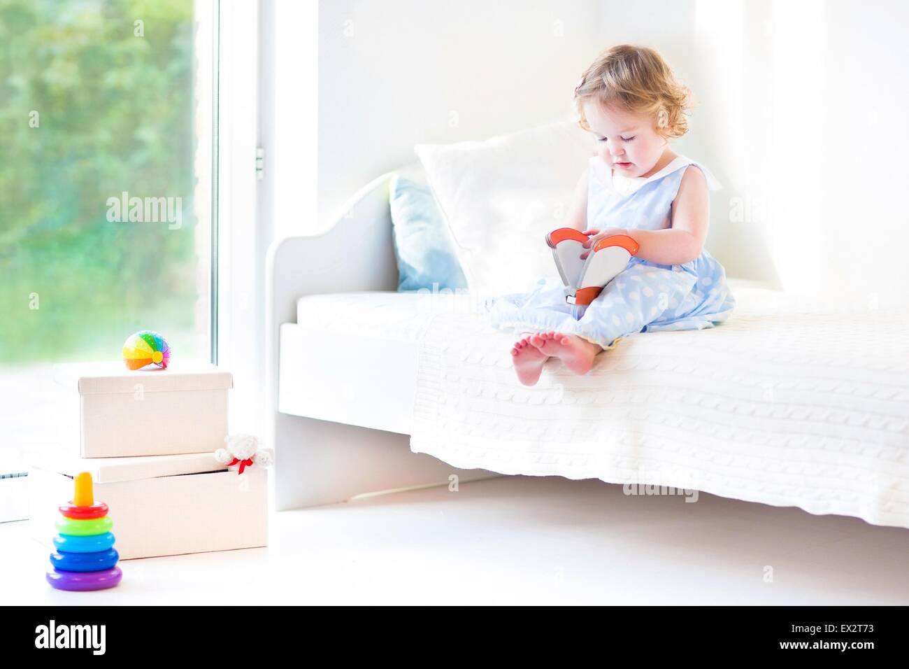 Beautiful curly toddler girl reading a book sitting on a white bed next to a big window with garden view Stock Photo