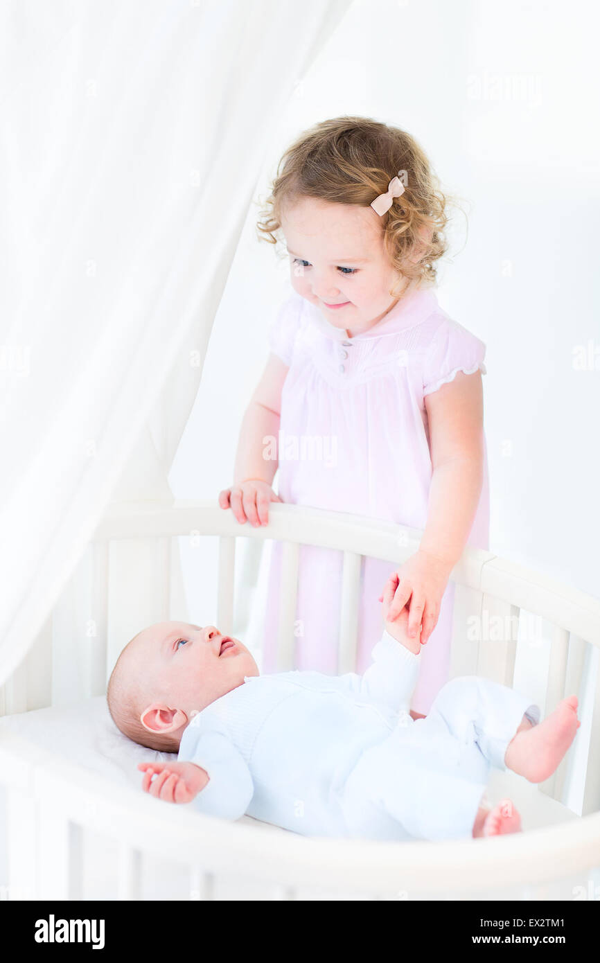 Happy toddler girl with curly hair wearing a pink dress holding her baby brother's hand in a white sunny bedroom Stock Photo