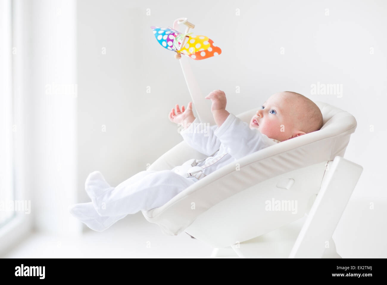 Cute newborn baby boy watching a colorful mobile toy sitting in a white high chair next to a window Stock Photo