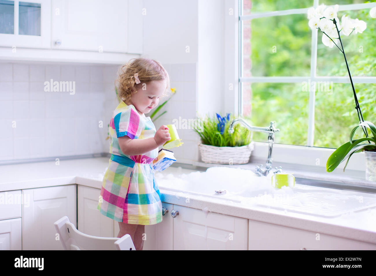 Child washing dishes. Kids wash plates and cups. Little girl helping in the kitchen playing with water and foam in a white sink  Stock Photo
