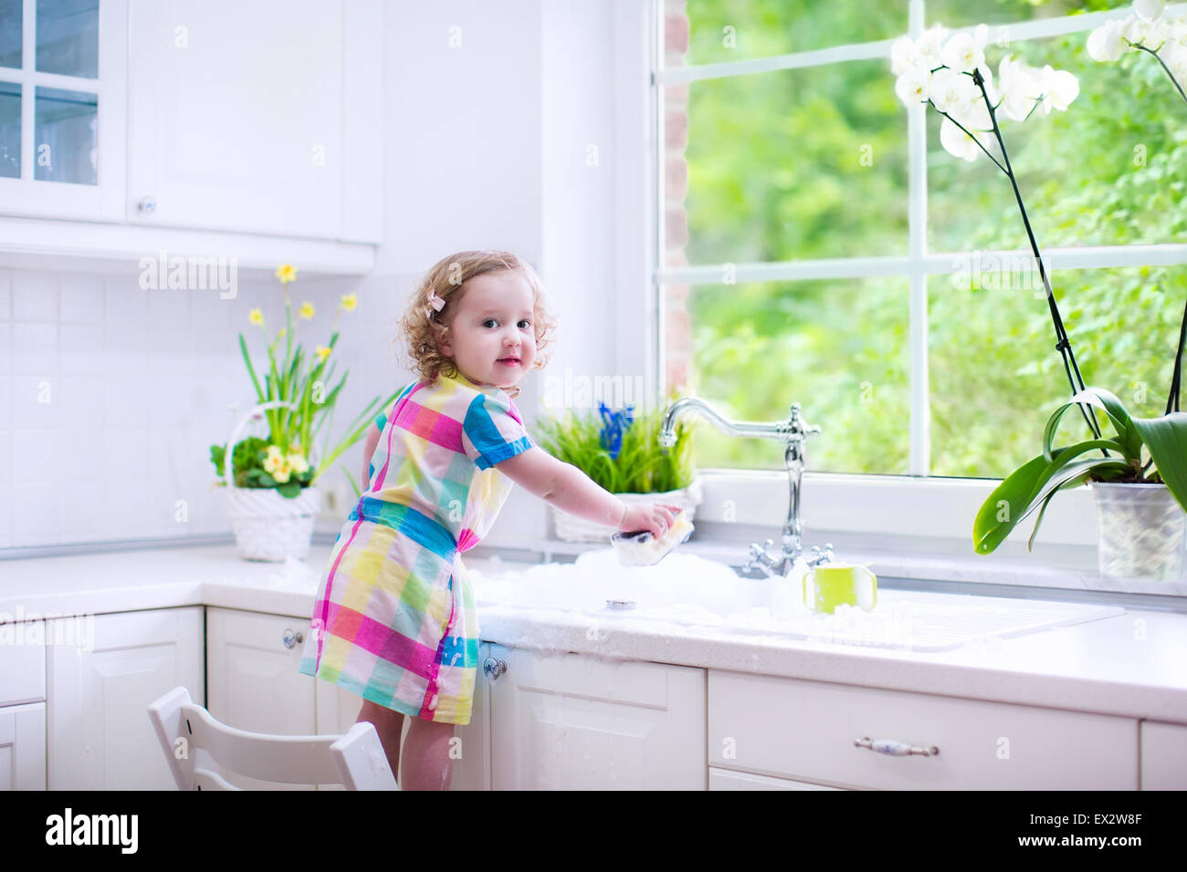 Child washing dishes. Kids wash plates and cups. Little girl helping in the kitchen playing with water and foam in a white sink Stock Photo