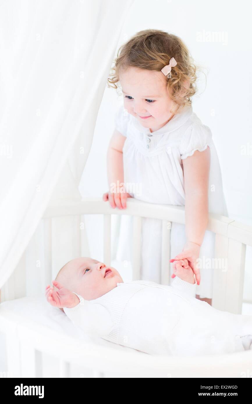 Happy toddler girl with curly hair wearing a pink dress holding her baby brother's hand in a white sunny bedroom Stock Photo