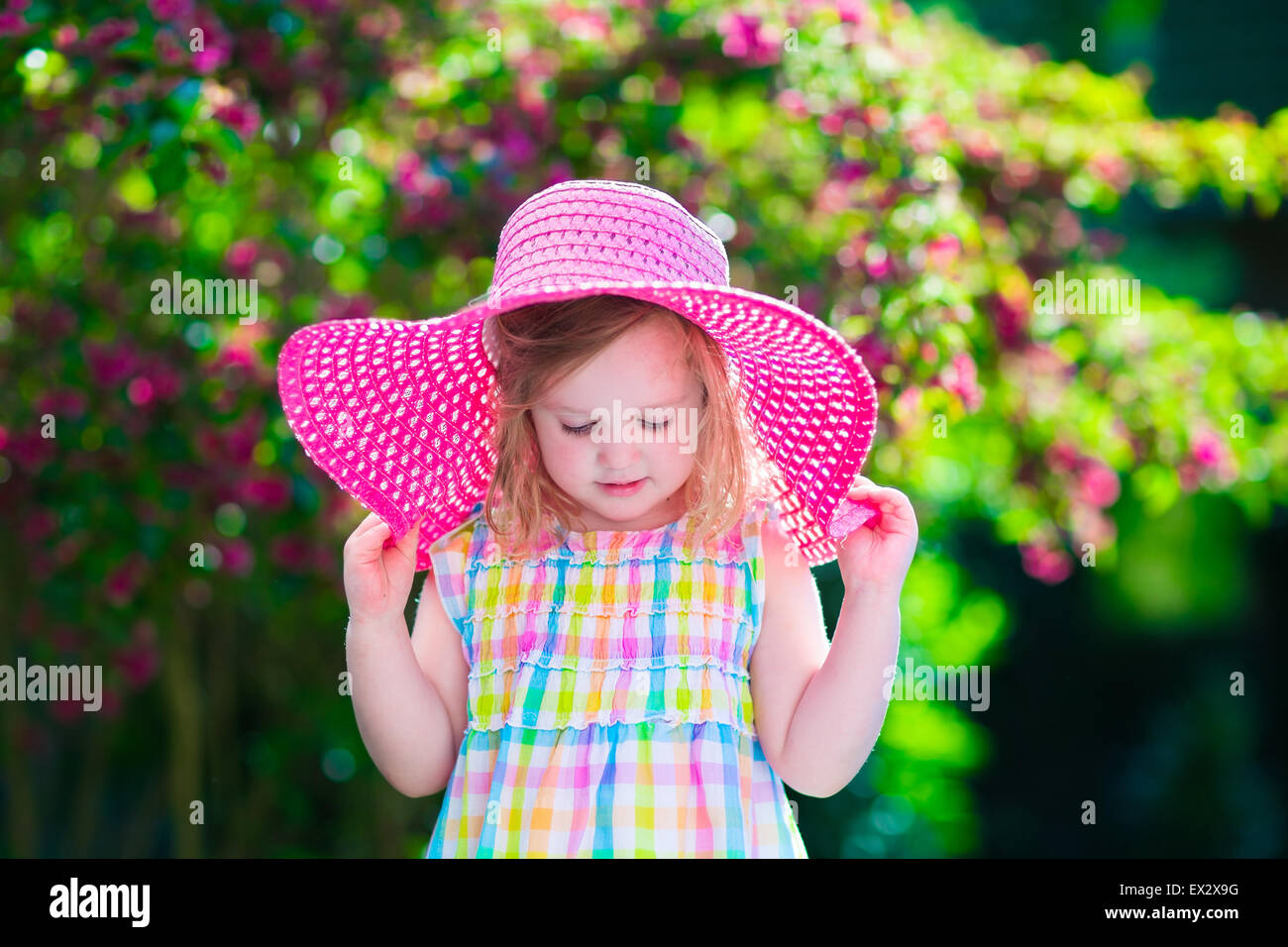 Little cute girl with flowers. Child wearing a pink hat playing in a blooming summer garden. Kids gardening. Stock Photo