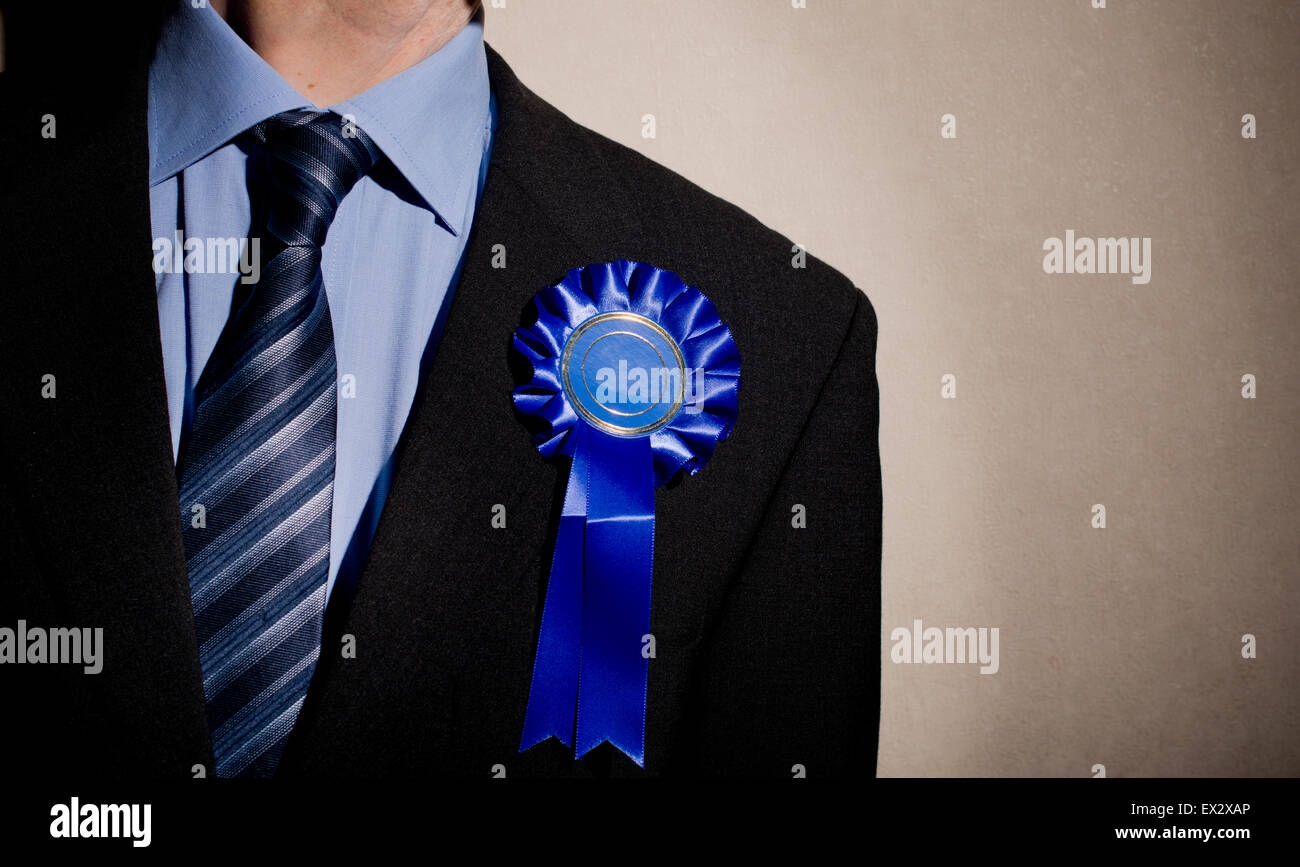 Election candidate wearing a dark business suit and a blue rosette., against a spot lit wall. Stock Photo