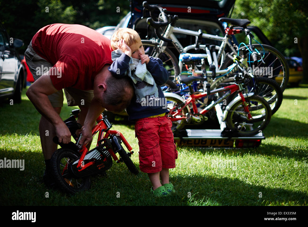 A young boy sucks his thumb and leans against his father who is mending the boy's bike before cycling Stock Photo
