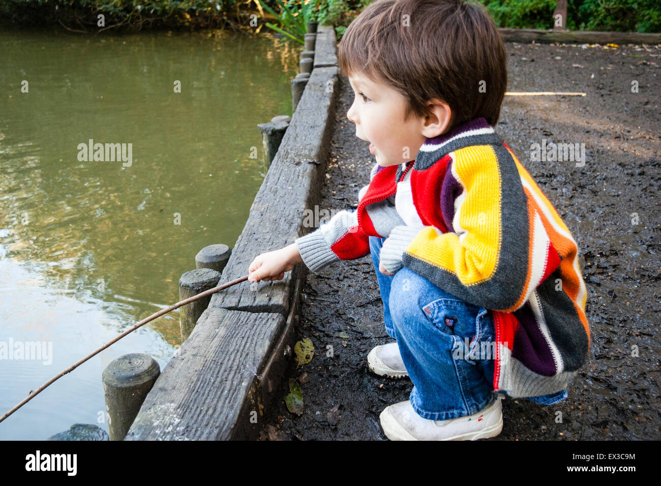 Child, boy, 4-5 year old, crouching down on dirt track by waterfront jetty  and holding stick as a fishing rod over still water of large pond Stock  Photo - Alamy