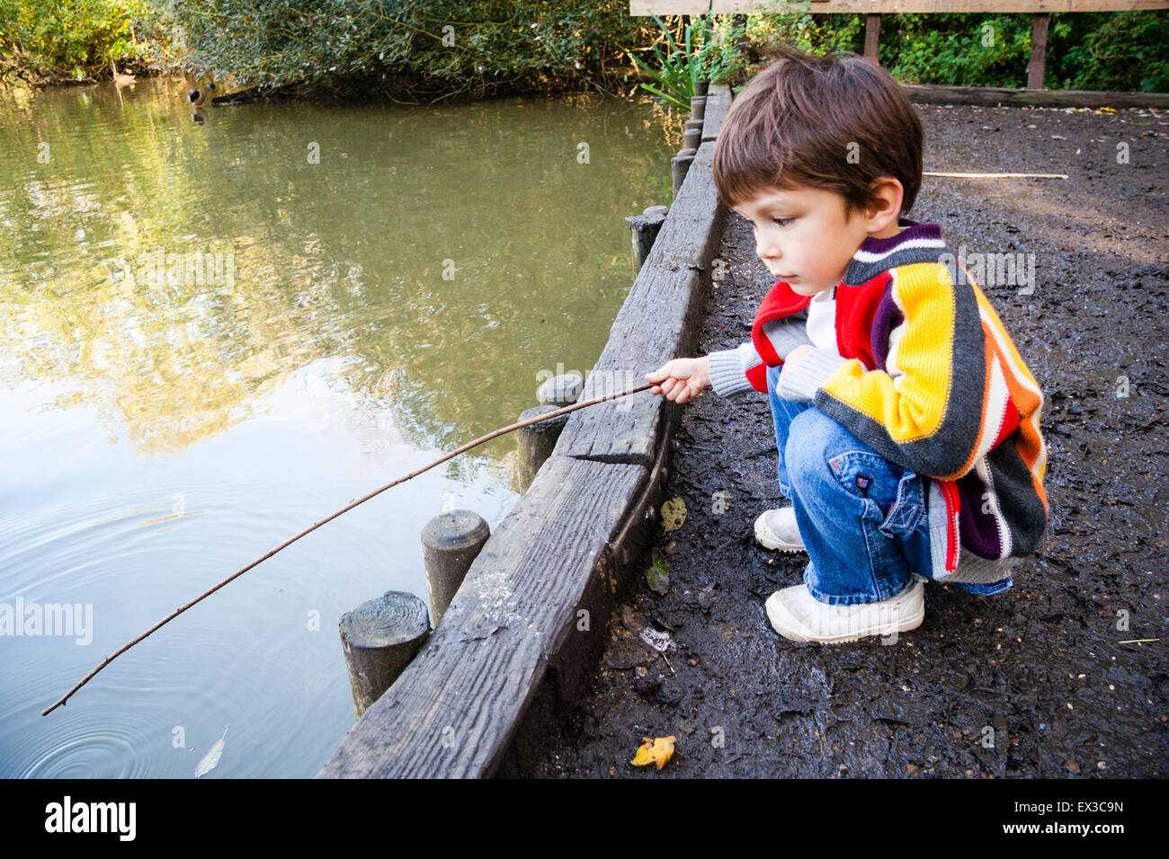 Child, boy, 4-5 year old, crouching down on dirt track by waterfront jetty  and holding stick as a fishing rod over still water of large pond Stock  Photo - Alamy