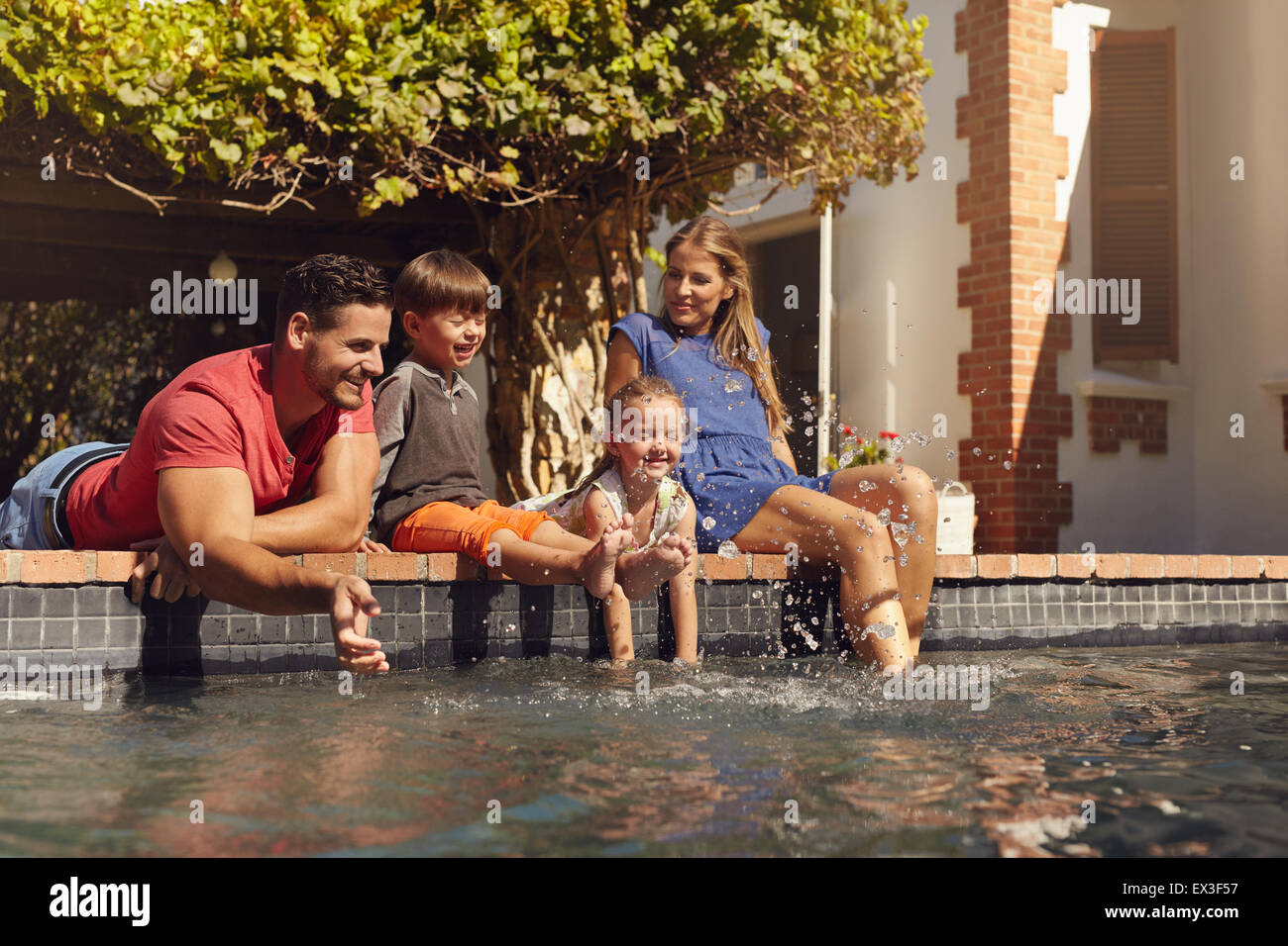 Outdoor shot of happy young family splashing water with hands and legs while sitting on edge of swimming pool. Family enjoying a Stock Photo