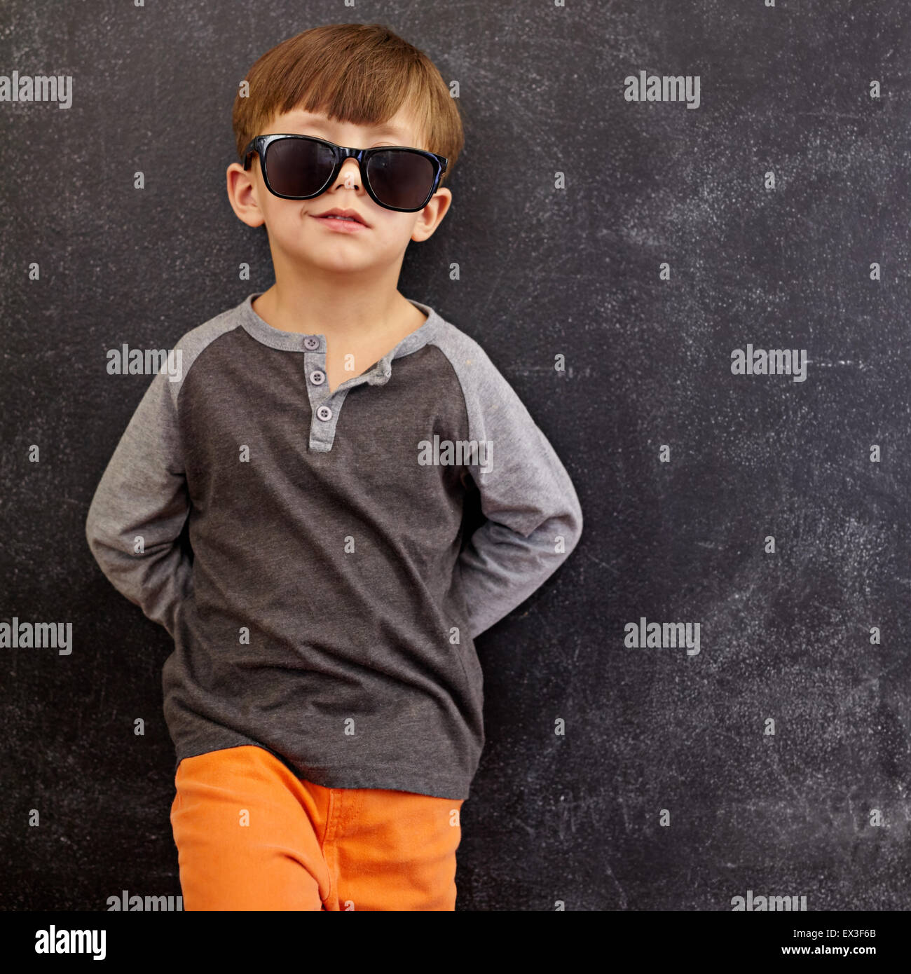 Portrait of smart little boy wearing sunglasses smirking. Cool kid in shades leaning on a blackboard looking at camera smiling w Stock Photo