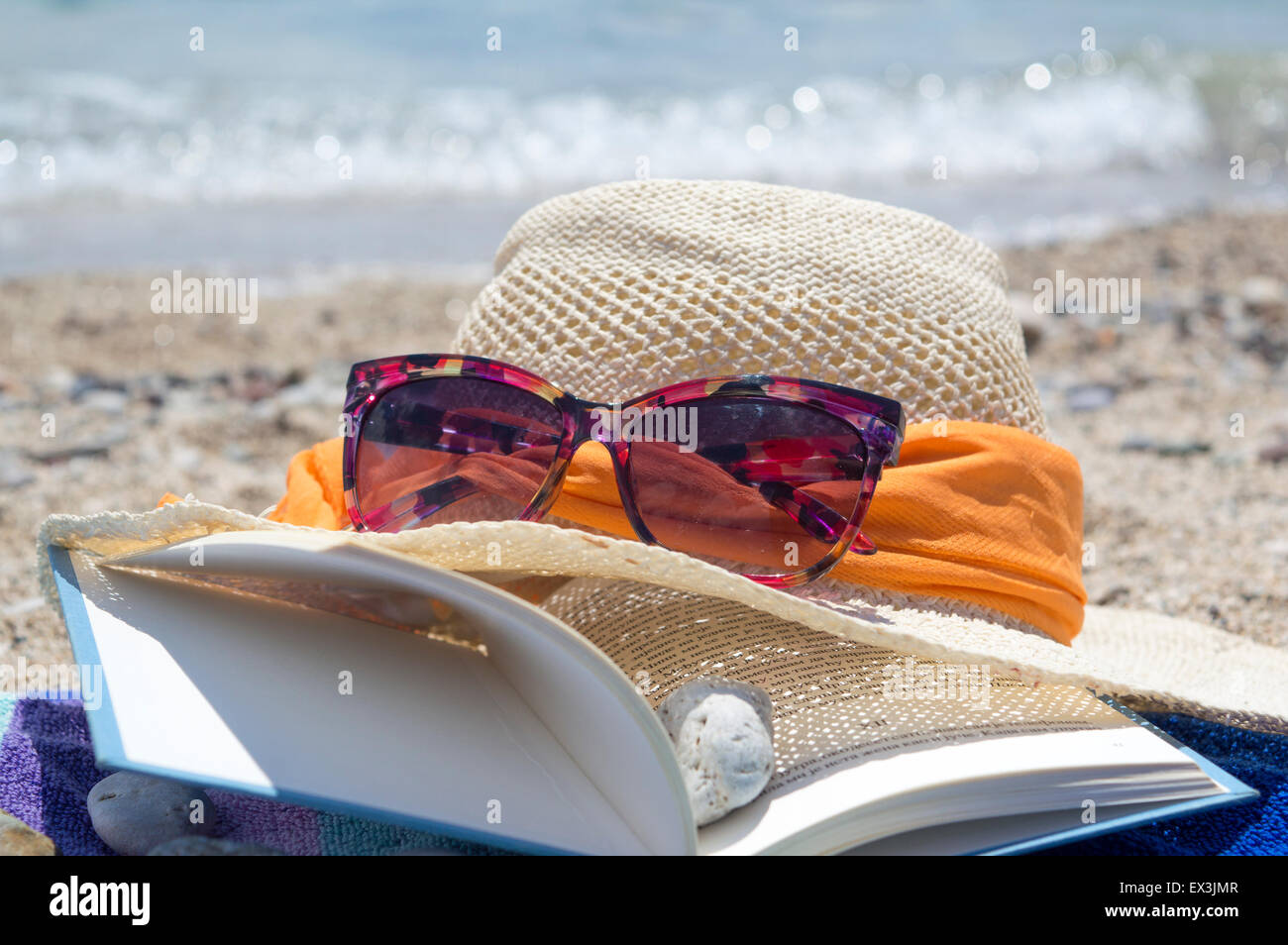 Straw hat sunglasses and a book on the beach with sea in backgound on a sunny day Stock Photo