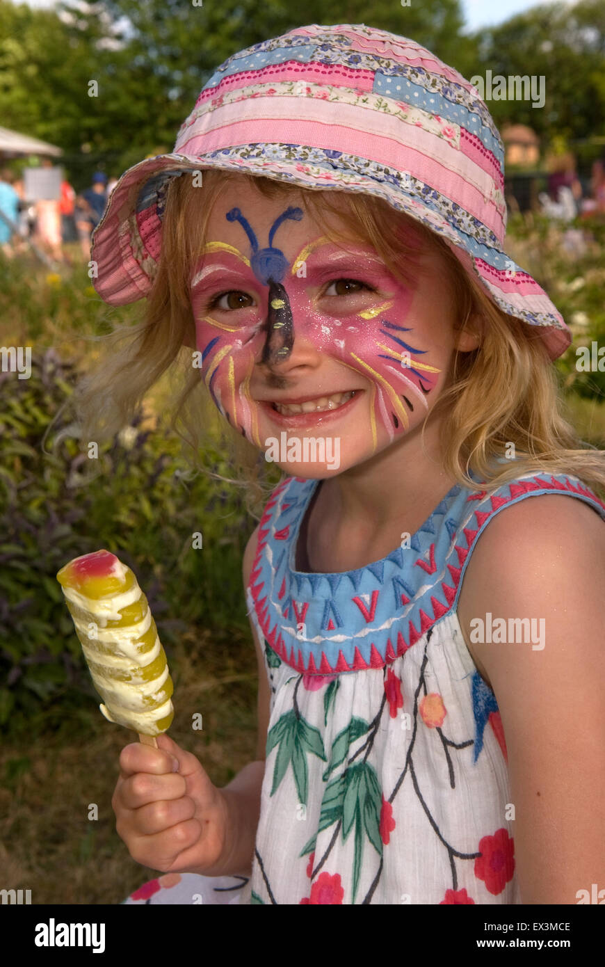 A happy, smiley 3 year old girl with painted face and ice lolly at a school summer fair, Headley, Hampshire, UK. Stock Photo