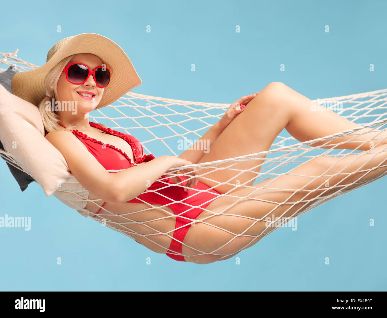 Beautiful woman in a red swimsuit wearing a stylish hat and lying in a hammock on blue background Stock Photo