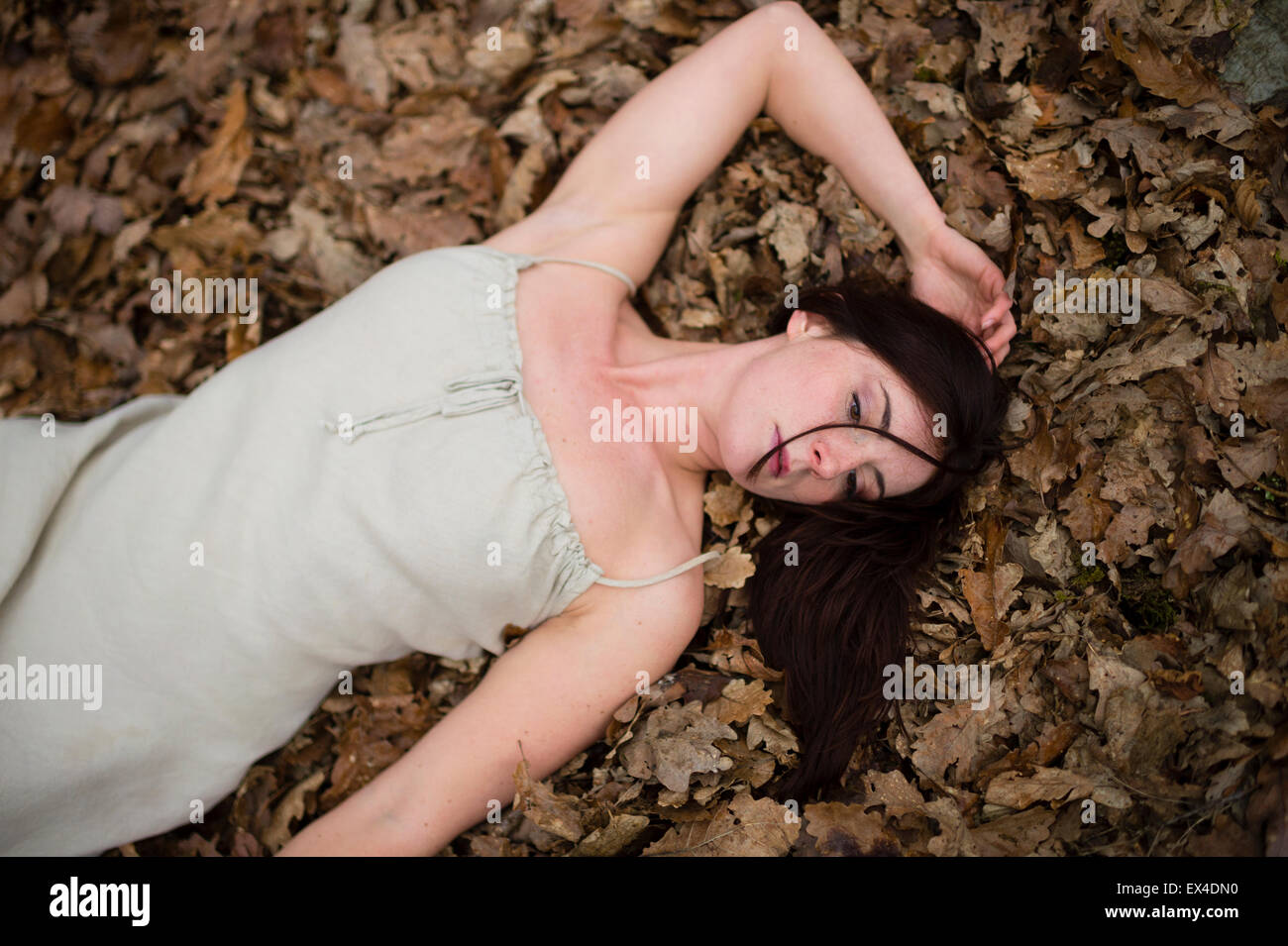 A young dark-haired caucasian woman girl  alone by herself in a bleak woodland forest lying on her back on the ground a bed of fallen leaves Stock Photo