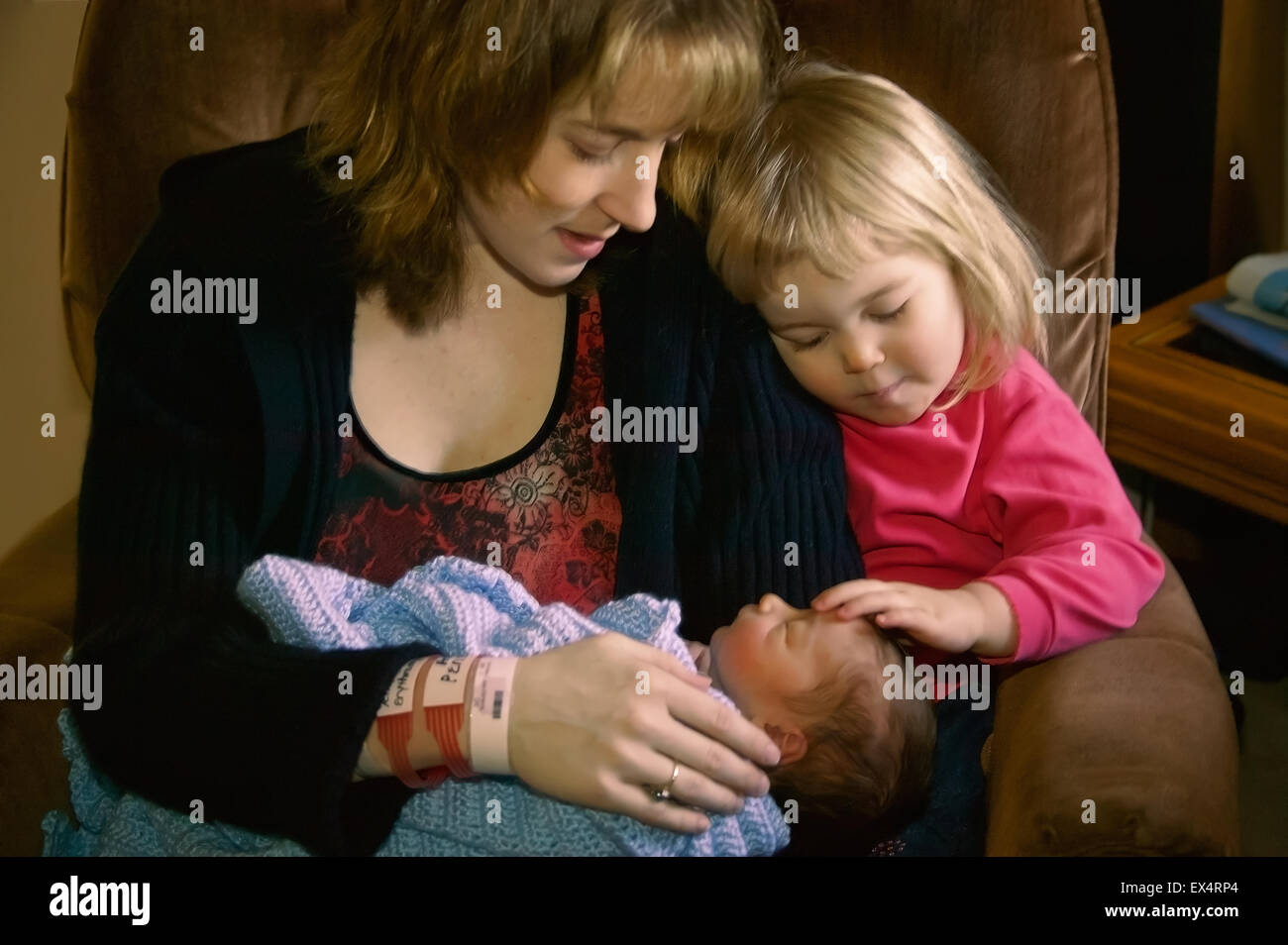 Mother with two year old daughter gently patting her day old baby brother on the head, after arriving home from the hospital Stock Photo