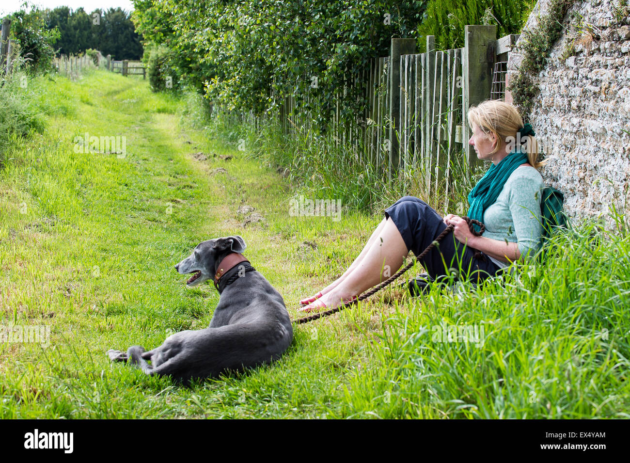 Resting during a dog walk.  Woman and greyhound sitting against a wall near a country footpath. Stock Photo