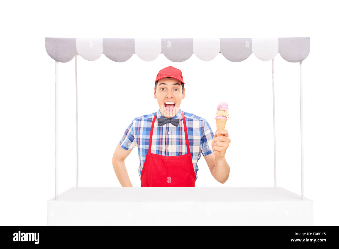 Excited young vendor with a red cap and apron holding an ice cream cone behind an ice cream stand isolated on white background Stock Photo