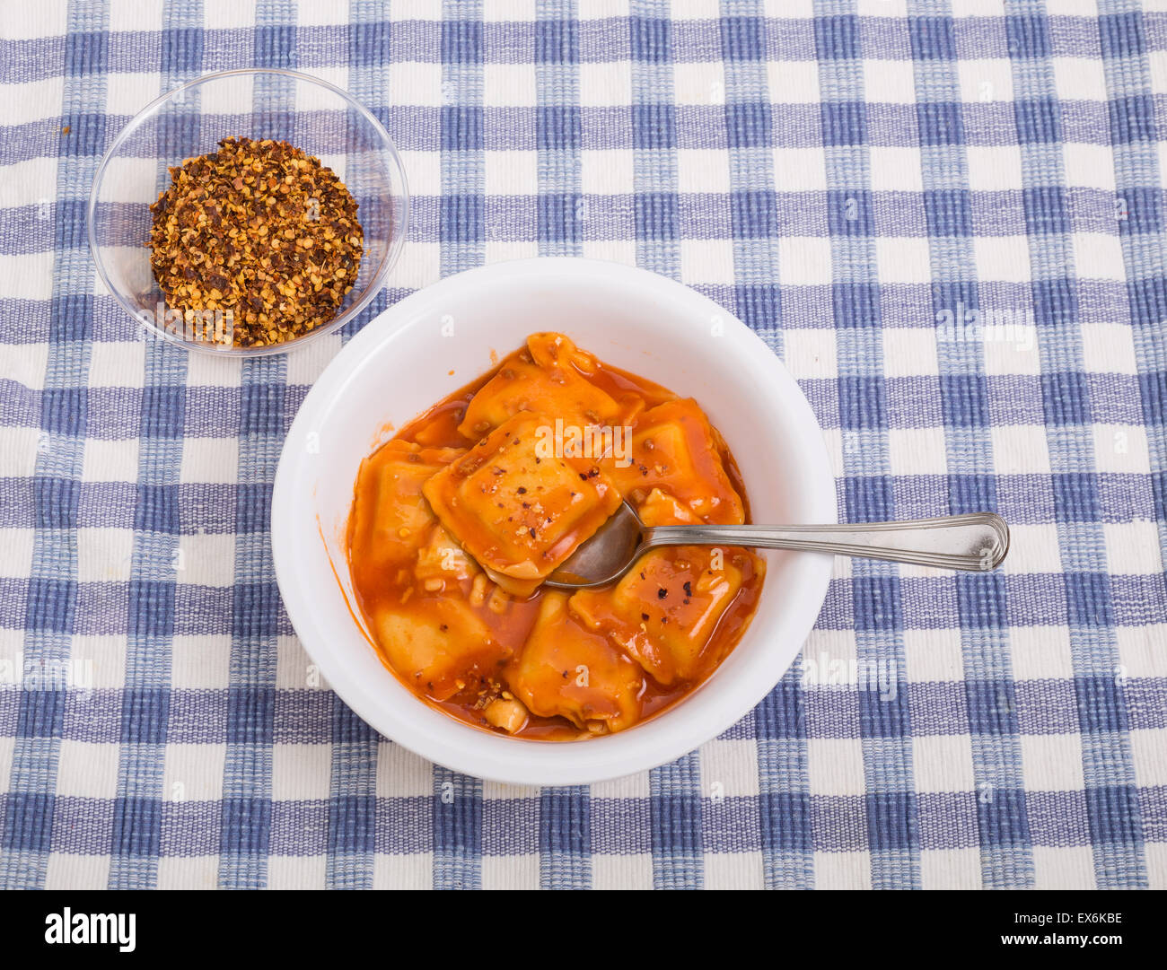 Canned ravioli in a white bowl on blue plaid tablecloth with red pepper ...