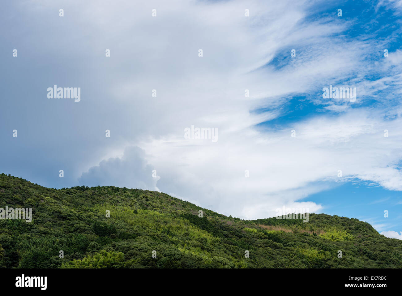 Dark storm clouds creeping in on an otherwise clear blue sky with a green tree filled mountain in the foreground. Stock Photo