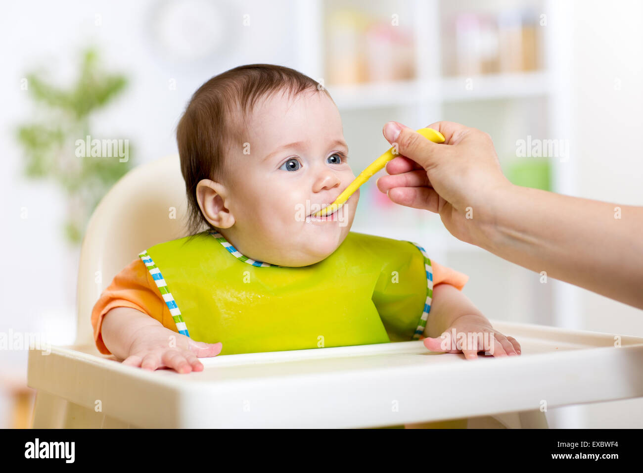 Mother feeding baby girl. Child sitting in hich chair in kitchen Stock