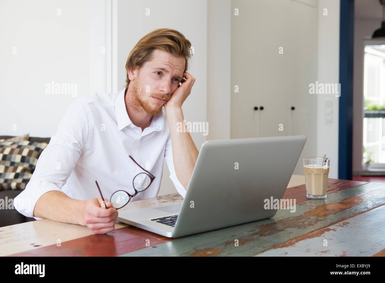 red-haired young man sitting at table with his laptop and looking sad Stock Photo