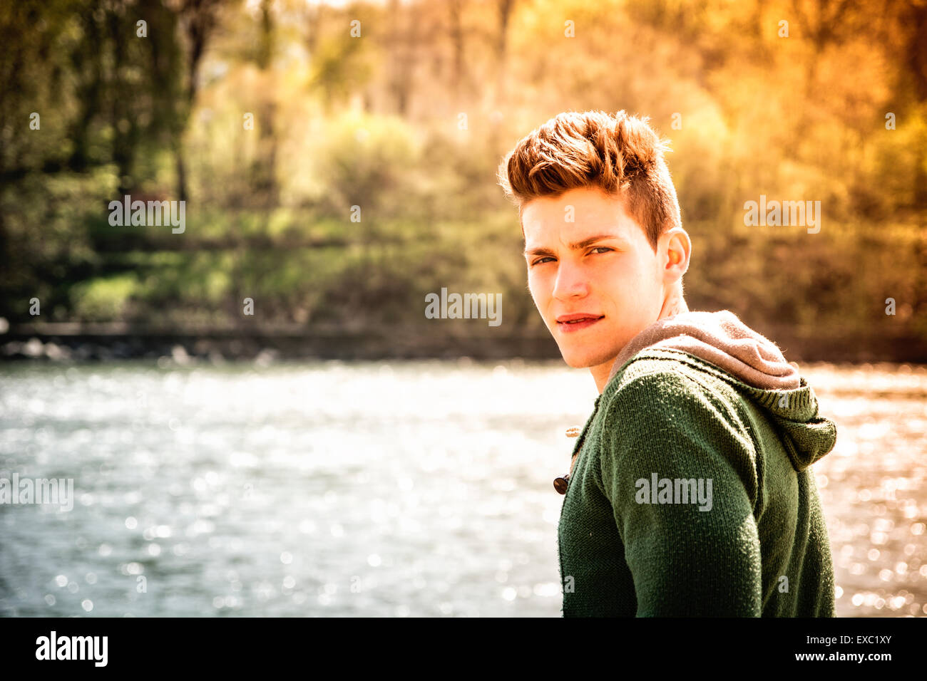 Head and shoulders length of contemplative light brown haired teenage boy wearing green hooded-shirt beside picturesque river or Stock Photo