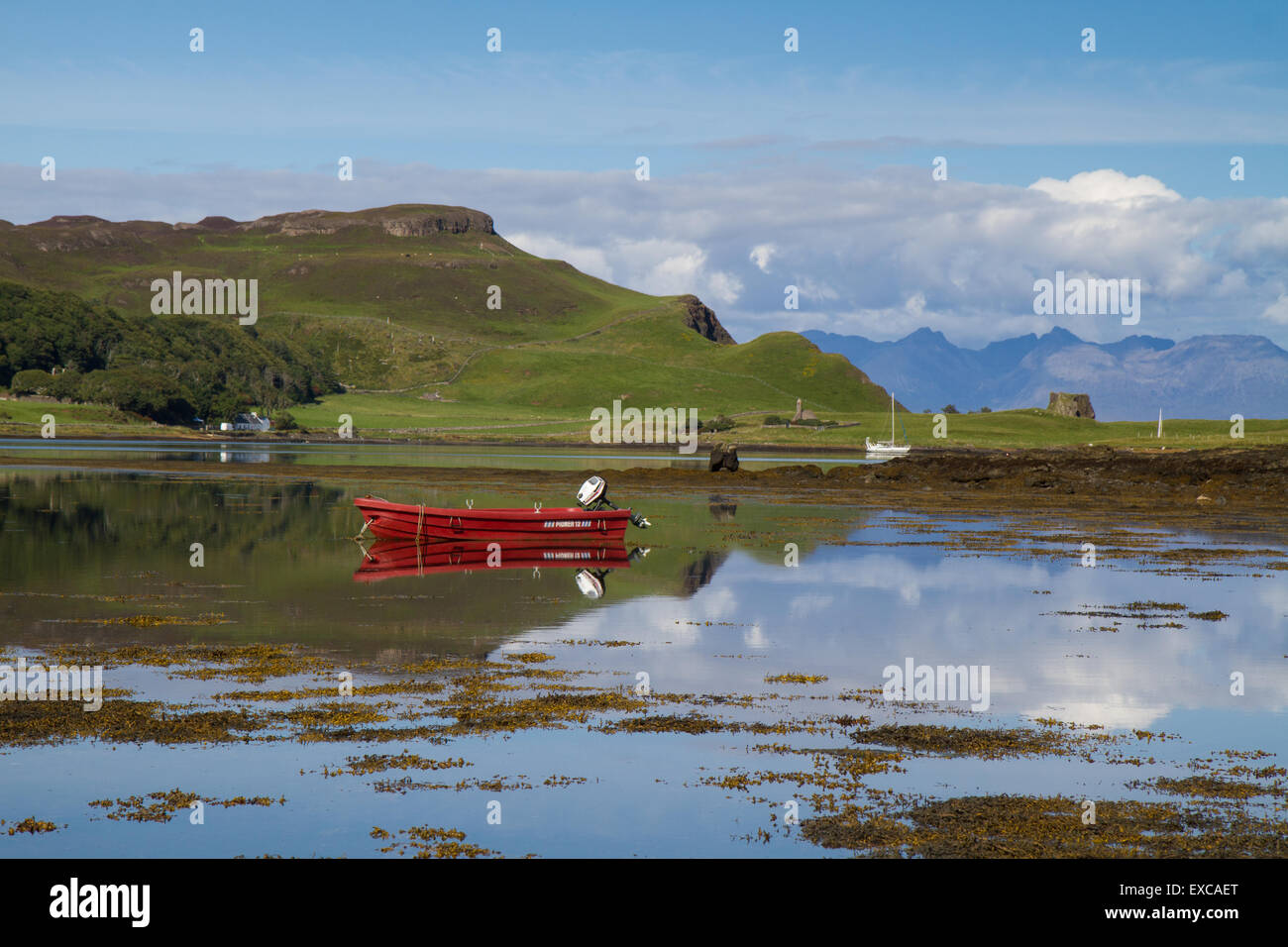 Small red boat reflected in a still natural harbor on the Isle of Canna with Rhum in the distance. Stock Photo