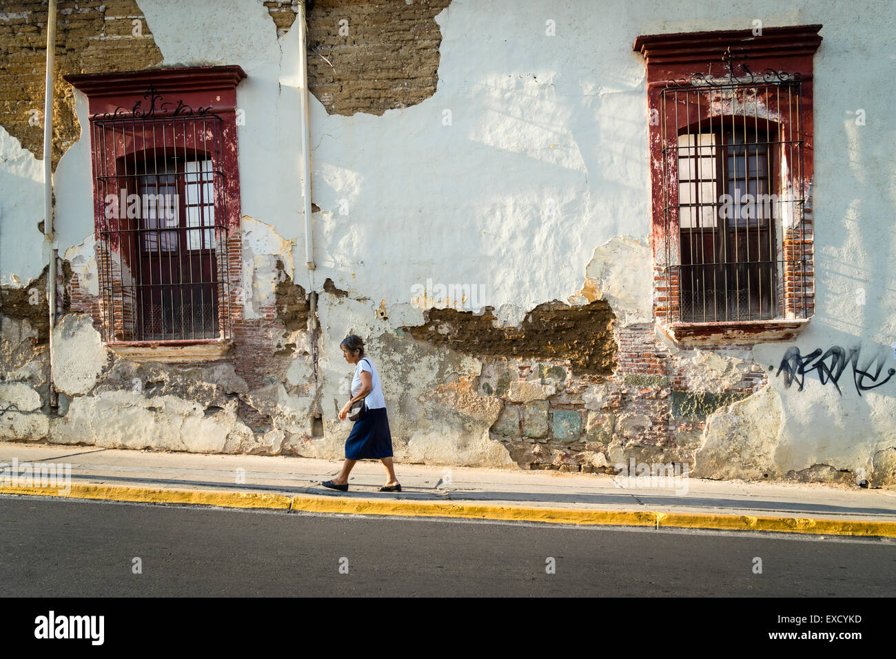 Woman walking up sloped street in front of old decaying stucco building facade Stock Photo