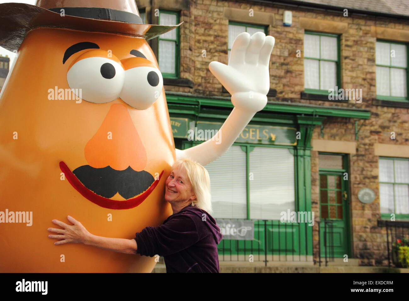 Belper, Derbyshire, UK. 12th July, 2015. Spud fan, Freda Raphael hugs a giant Mr Potato Head that has re-appeared in a Derbyshire town. The 7ft-tall fibreglass statue was gifted to Belper in 2001 by its twin town, Pawtucket, Rhode Island, USA. After being dubbed a 'monstrosity' by some locals, the divisive character made national headlines. It was vandalised and banished. Now the spud has been spruced up by a local youth group and unveiled at Belper’s food festival today. Credit:  Deborah Vernon/Alamy Live News Stock Photo