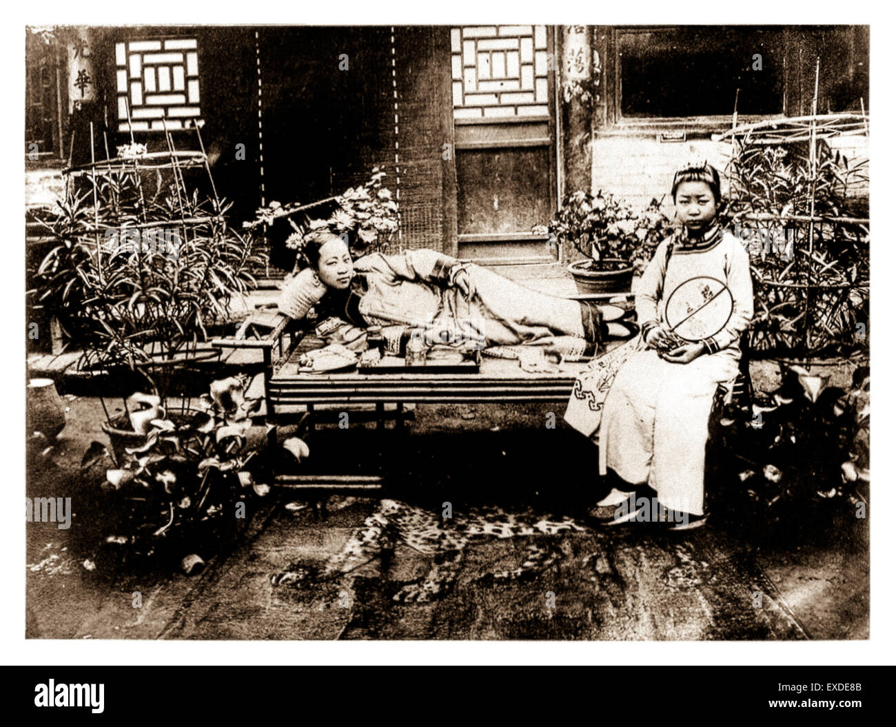 'A Manchurian lady smoking opium', taken before 1909.  A wealthy lady reclining on an opium bed in a courtyard holds an opium pipe and 'yen hock' (opium needle). In front of her is a tray holding the other paraphernalia associated with opium smoking. To her left an attendant holding a fan. See description for more information. Stock Photo