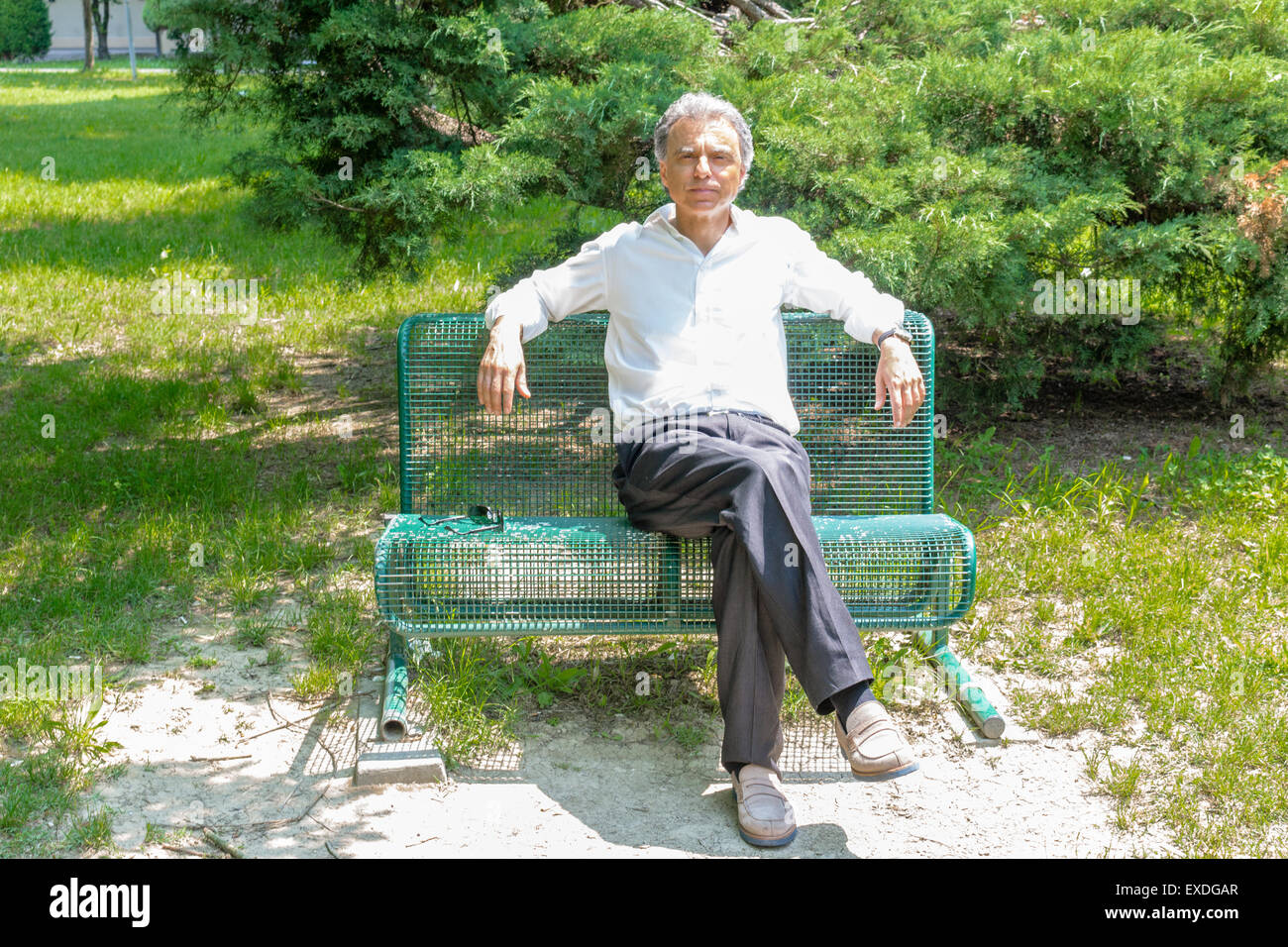 Handsome middle-aged man with salt pepper hair dressed with white shirt, blue slacks and beige moccasins is resting on a bench in city park keeping his arms opened: he shows a reassuring look Stock Photo