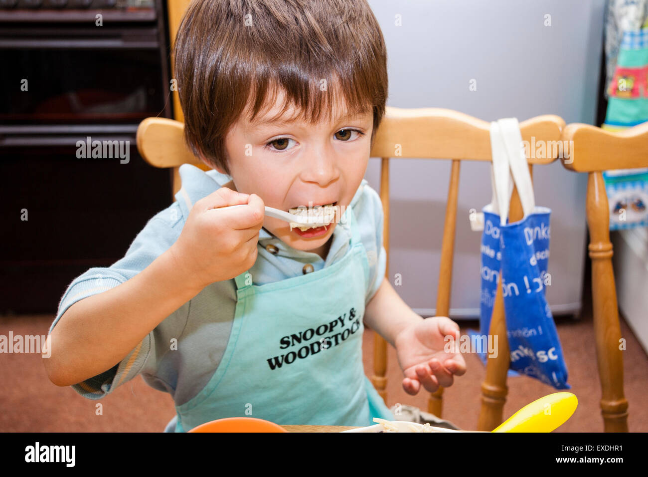 Caucasian child, boy, 4-5 year old, sitting indoors at the dinner table stuffing a spoonful of pasta into his mouth. Wears a cooking apron. Stock Photo
