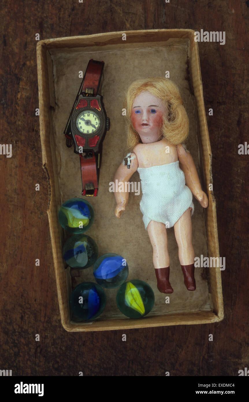 Small antique bisque doll with brown boots and blonde hair lying in box with tin toy watch and glass marbles Stock Photo