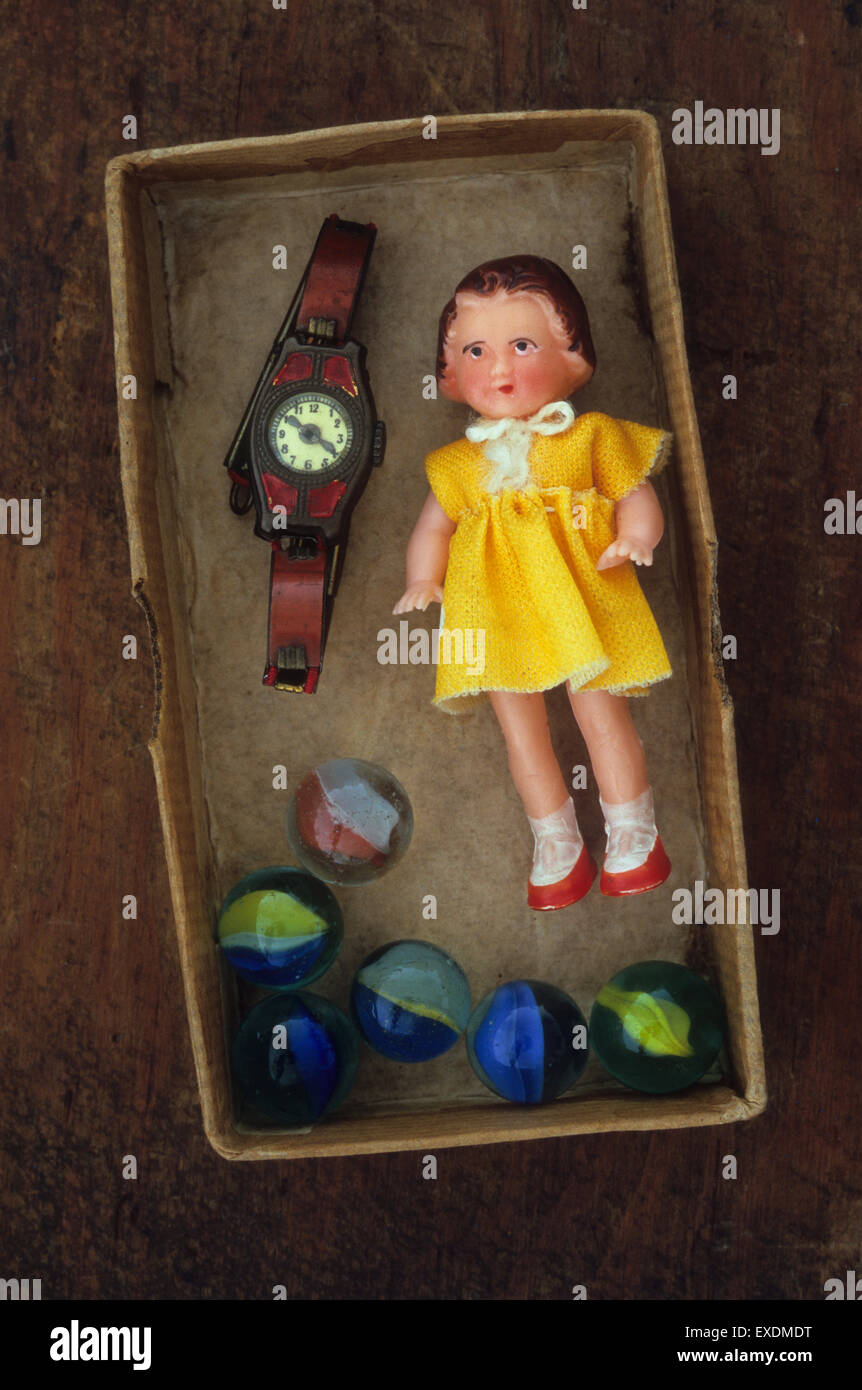 Small plastic vintage doll in yellow dress lying  in cardboard box with tin toy watch and glass marbles Stock Photo