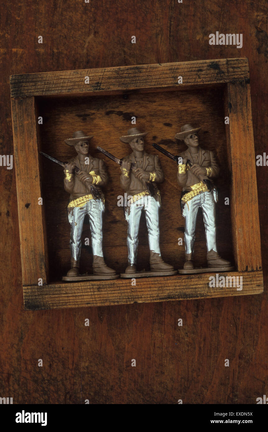 Three identical plastic models of cowboys with silver trousers holding rifles over shoulders standing in wooden box Stock Photo