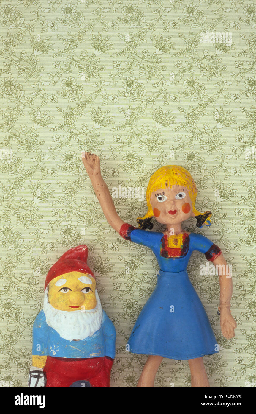 Bendy rubber girl doll with blue dress standing with arm raised next to gnome in front of floral wallpaper Stock Photo