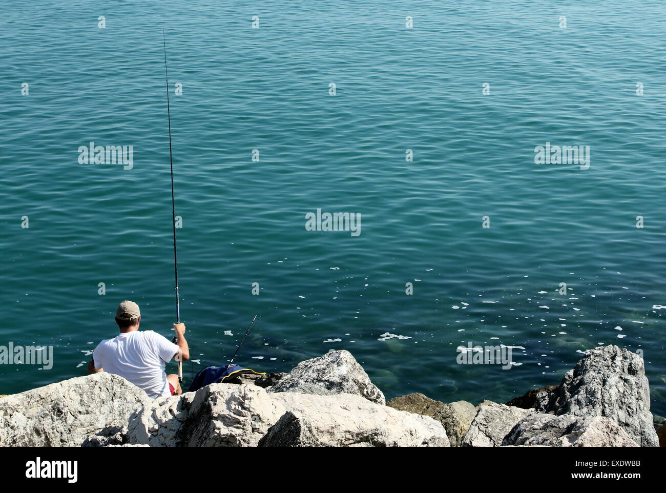 A man, a fisherman, is above the large rock placed near the sea. Stock Photo