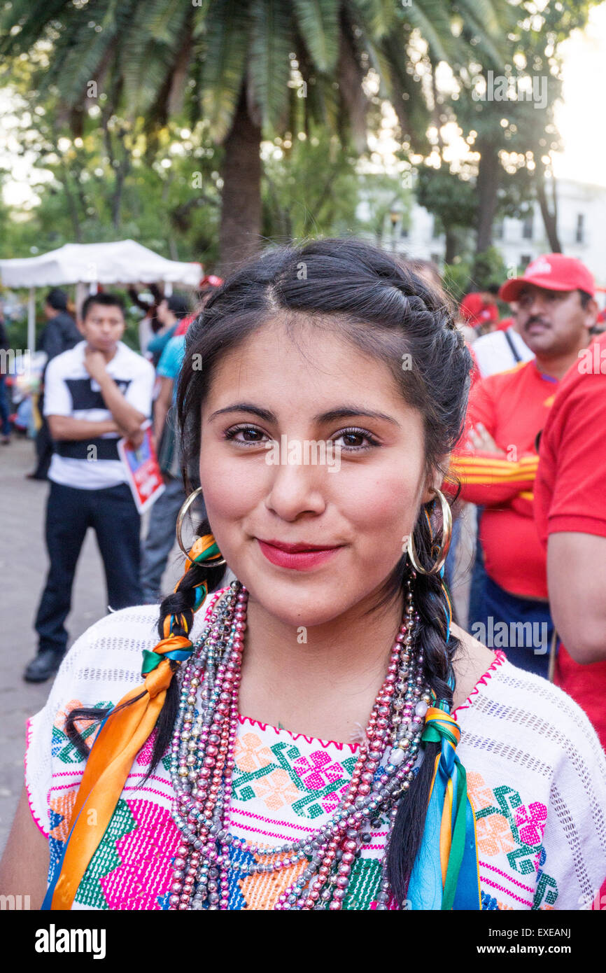 pretty Mexican girl with ribbons woven into her braids & wearing traditional costume stands out in crowd of red shirts at rally Stock Photo