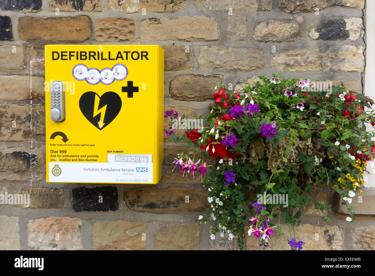 Heart defibrillator life saving equipment for cardiac arrest with code lock for public use in a town with flower basket  England Stock Photo
